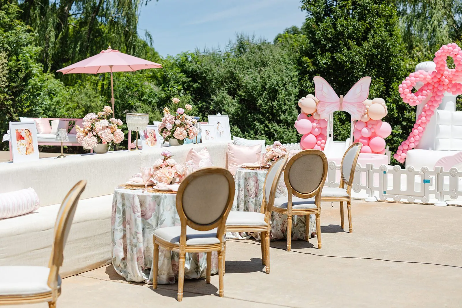 A garden celebration styled by Adorn Events — floral-draped tables, a blush parasol, and a butterfly balloon arch beneath the trees